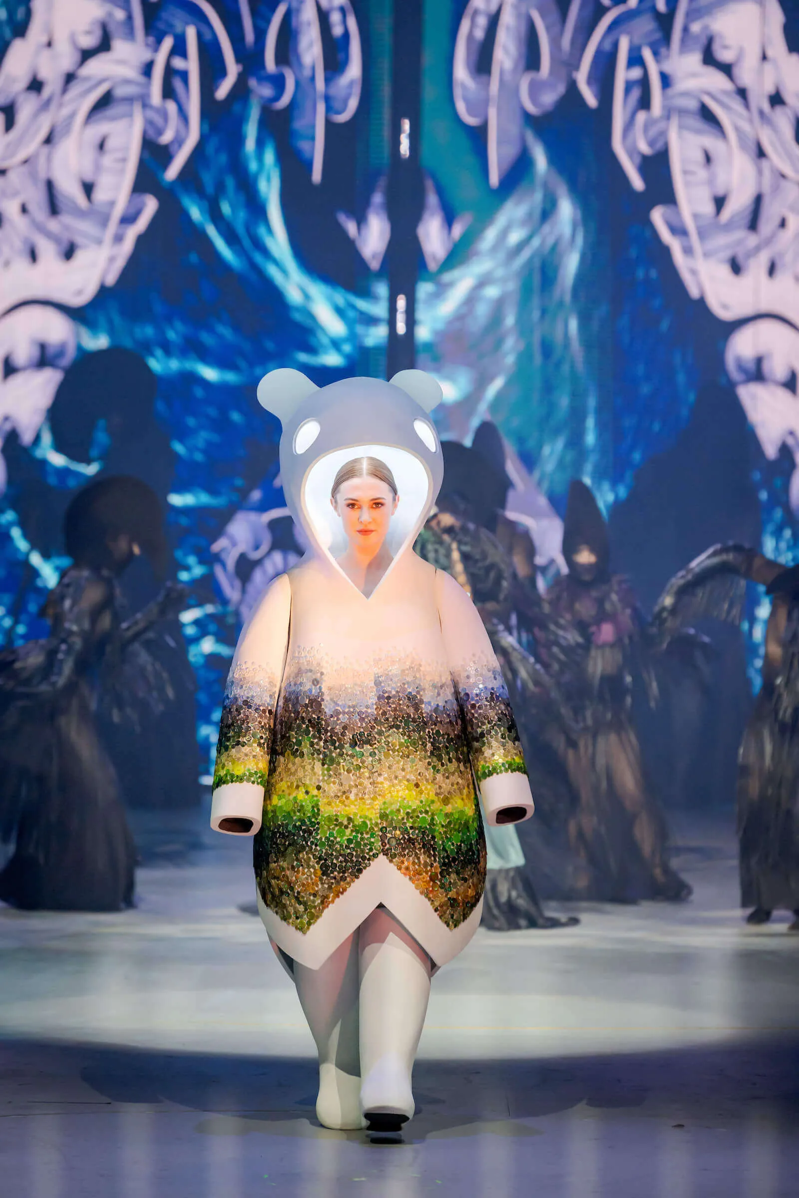 Model wearing a white bear-themed costume with colorful abstract patterns walking on a runway with blue artistic background and performers in black costumes.
